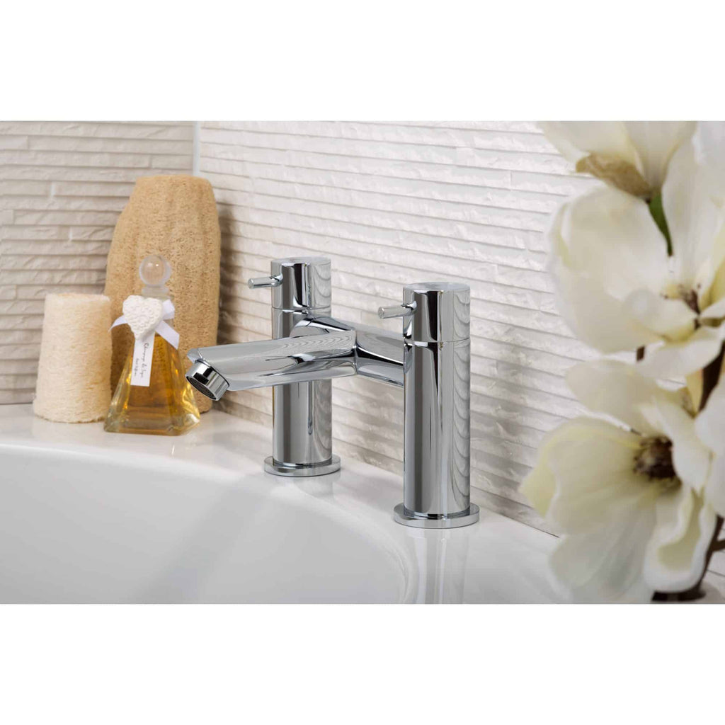 Sleek chrome bathroom tap with dual handles on a modern washbasin, surrounded by beige towels, a decorative soap dispenser, and white orchids, showcasing elegant bathroom fixtures and design.