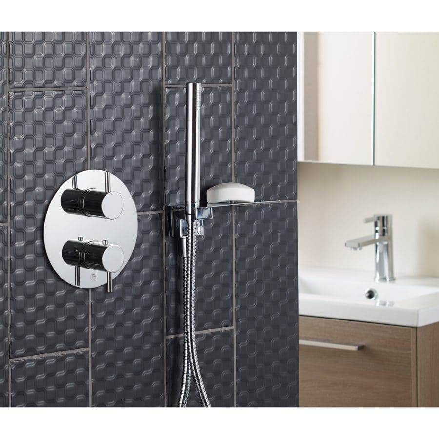 Shower tap control on black geometric tiled wall with shower head and bathroom sink in the background.