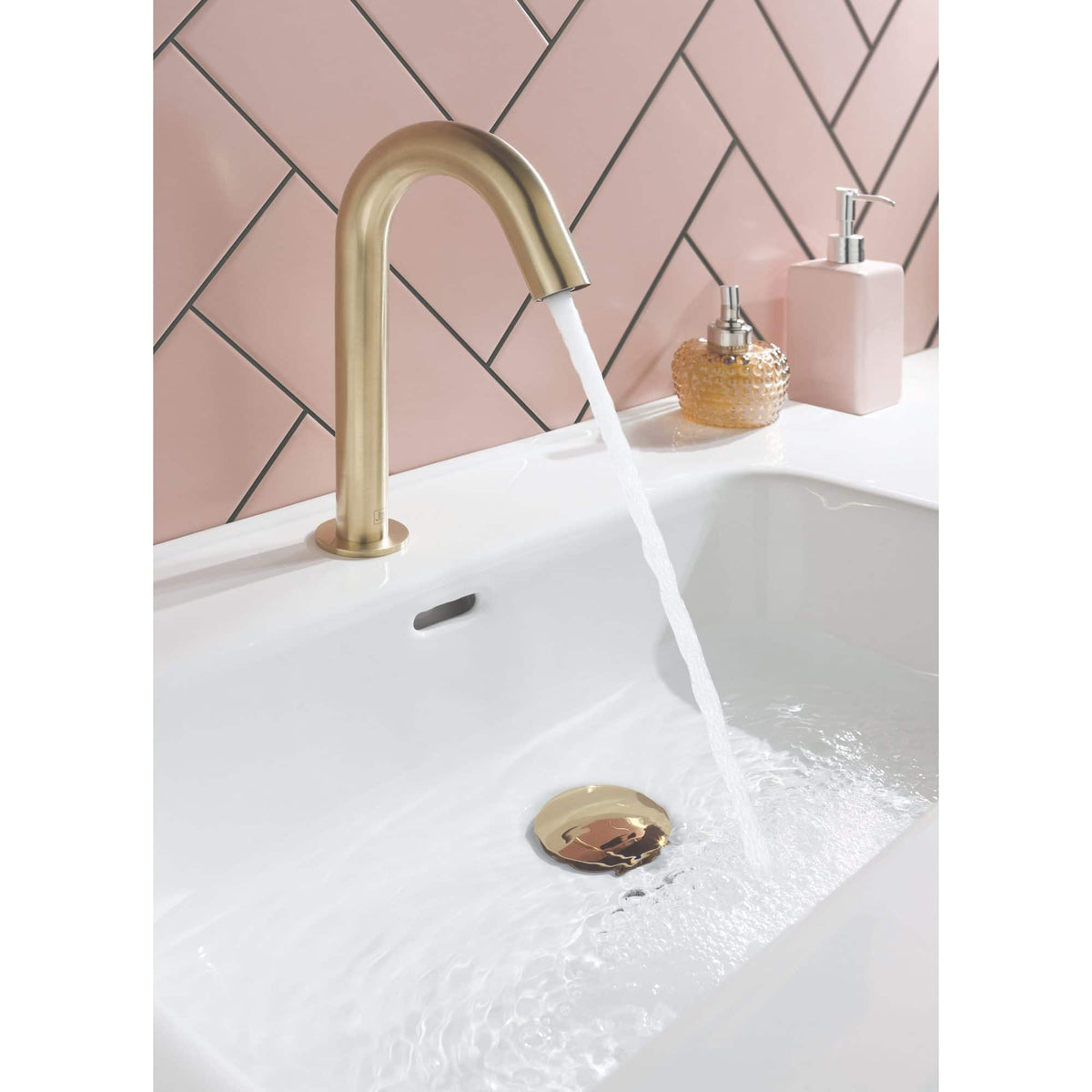 Sleek gold bathroom sink tap with running water, modern design, pink tiled wall, and stylish bathroom accessories for Bubly Bathrooms in the UK.
