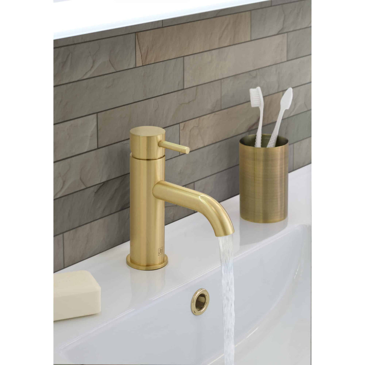 Polished gold bathroom faucet with running water, matching toothbrush holder with toothbrushes, set against a modern tiled wall for luxury bathroom decor.