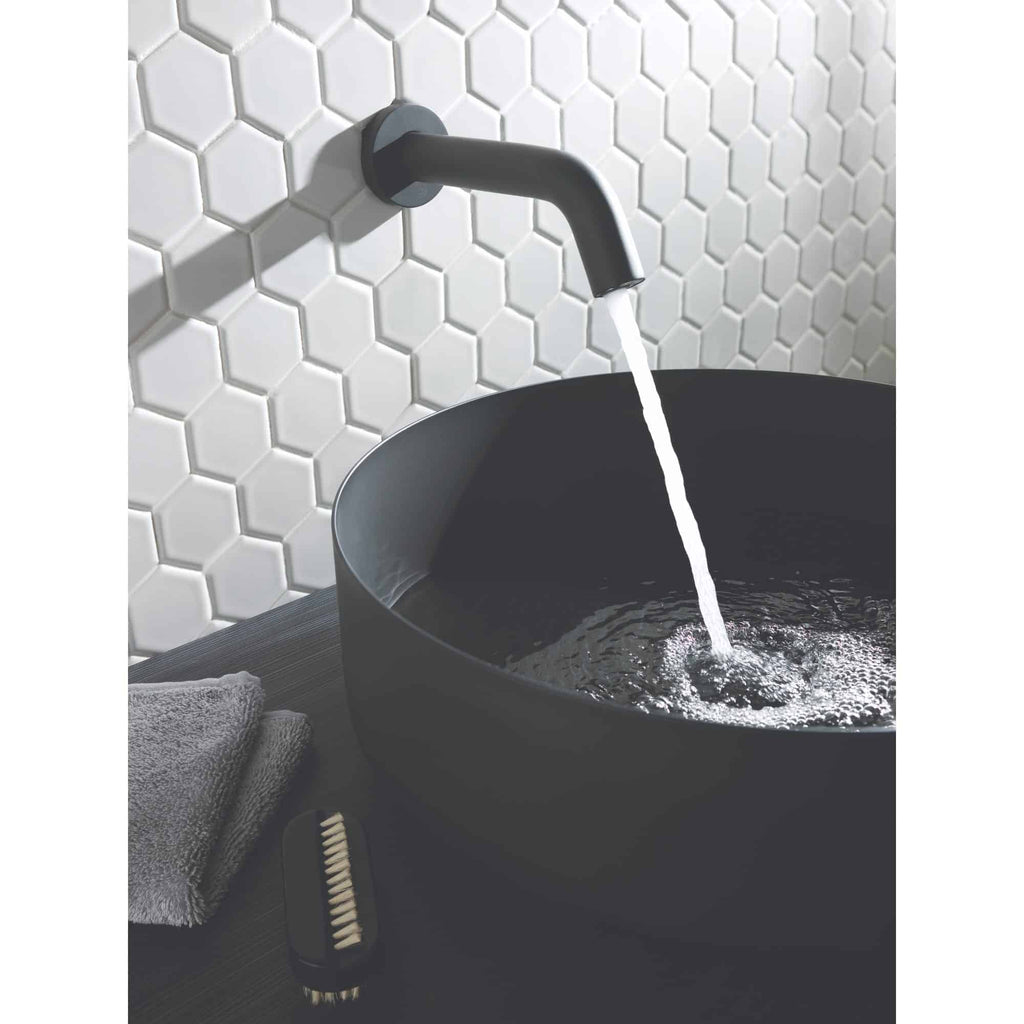 Black bathroom sink with wall-mounted faucet and running water, modern bathroom design with white hexagonal tiled wall, grey towel, and cleaning brush, exemplifying stylish and functional bathroom fixtures.