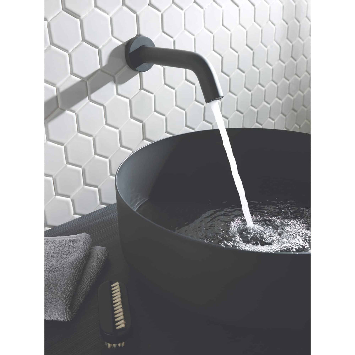 Black bathroom sink with wall-mounted faucet and running water, modern bathroom design with white hexagonal tiled wall, grey towel, and cleaning brush, exemplifying stylish and functional bathroom fixtures.