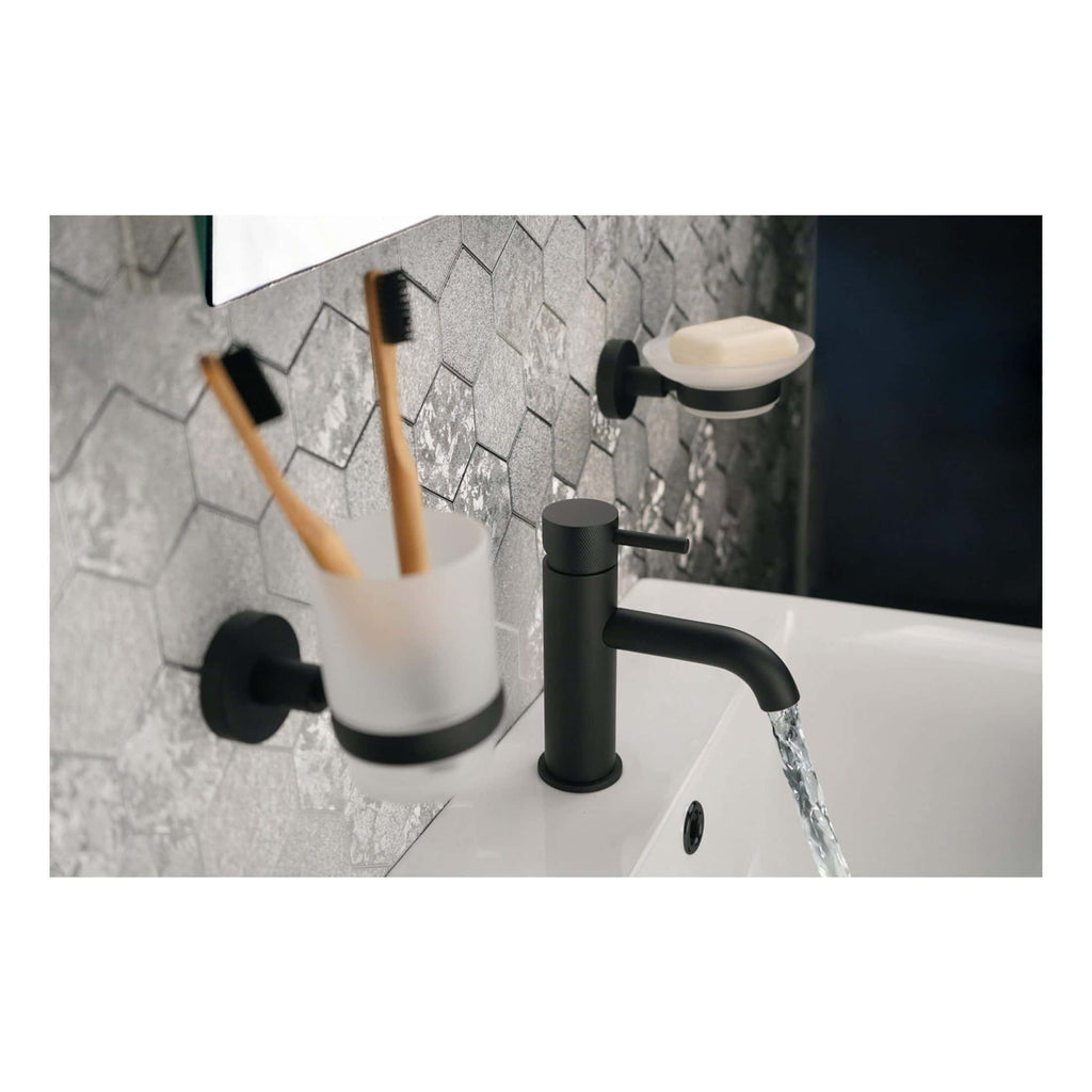 Sleek black bathroom tap with water flowing into a white basin, surrounded by modern bathroom accessories including toothbrush holders and soap dishes, featuring textured grey hexagonal tiled wall for sophisticated bathroom design.