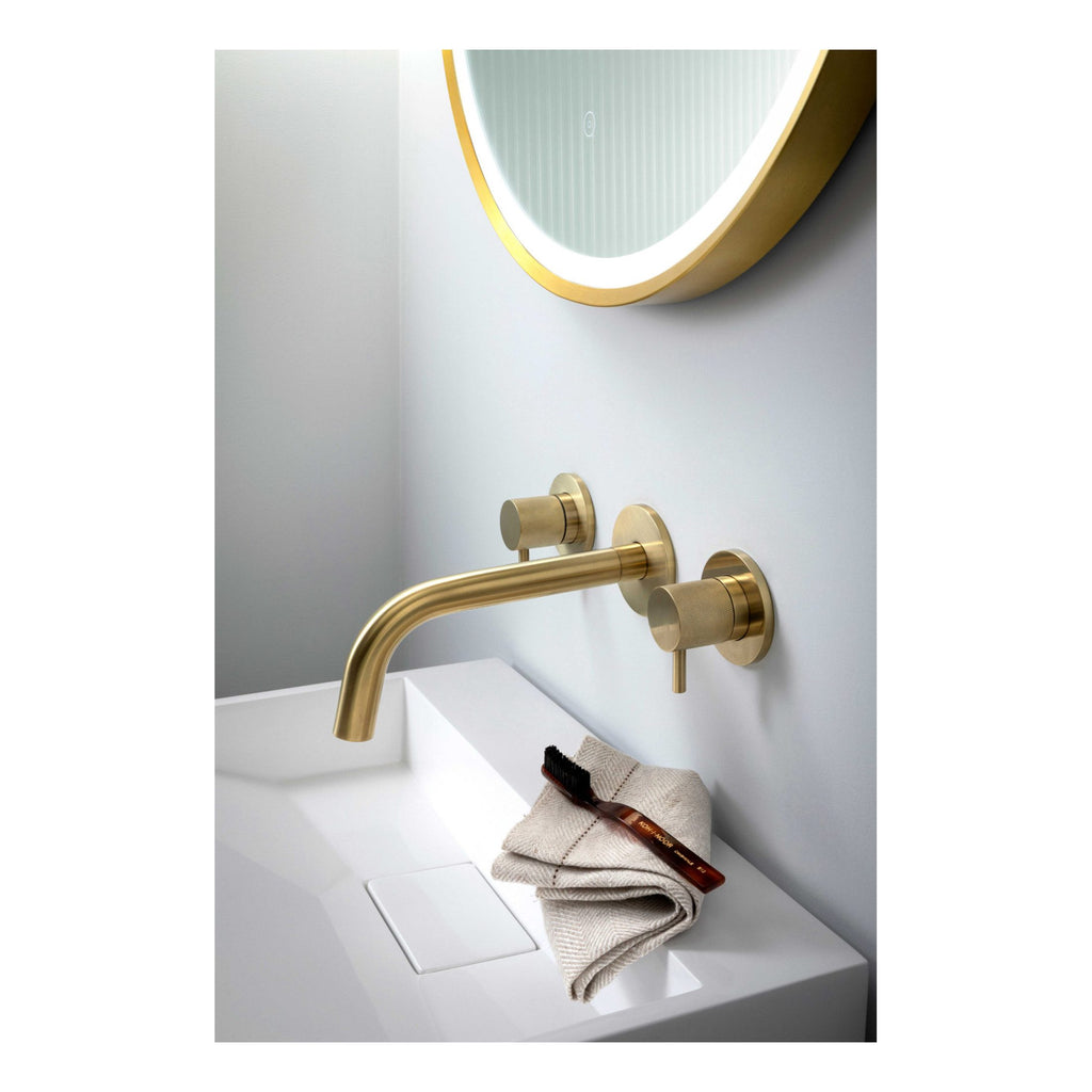Sleek gold bathroom faucet with wall-mounted controls above a modern white sink, complemented by round illuminated mirror, showcasing elegant bathroom fixtures for luxurious Bubly Bathrooms.