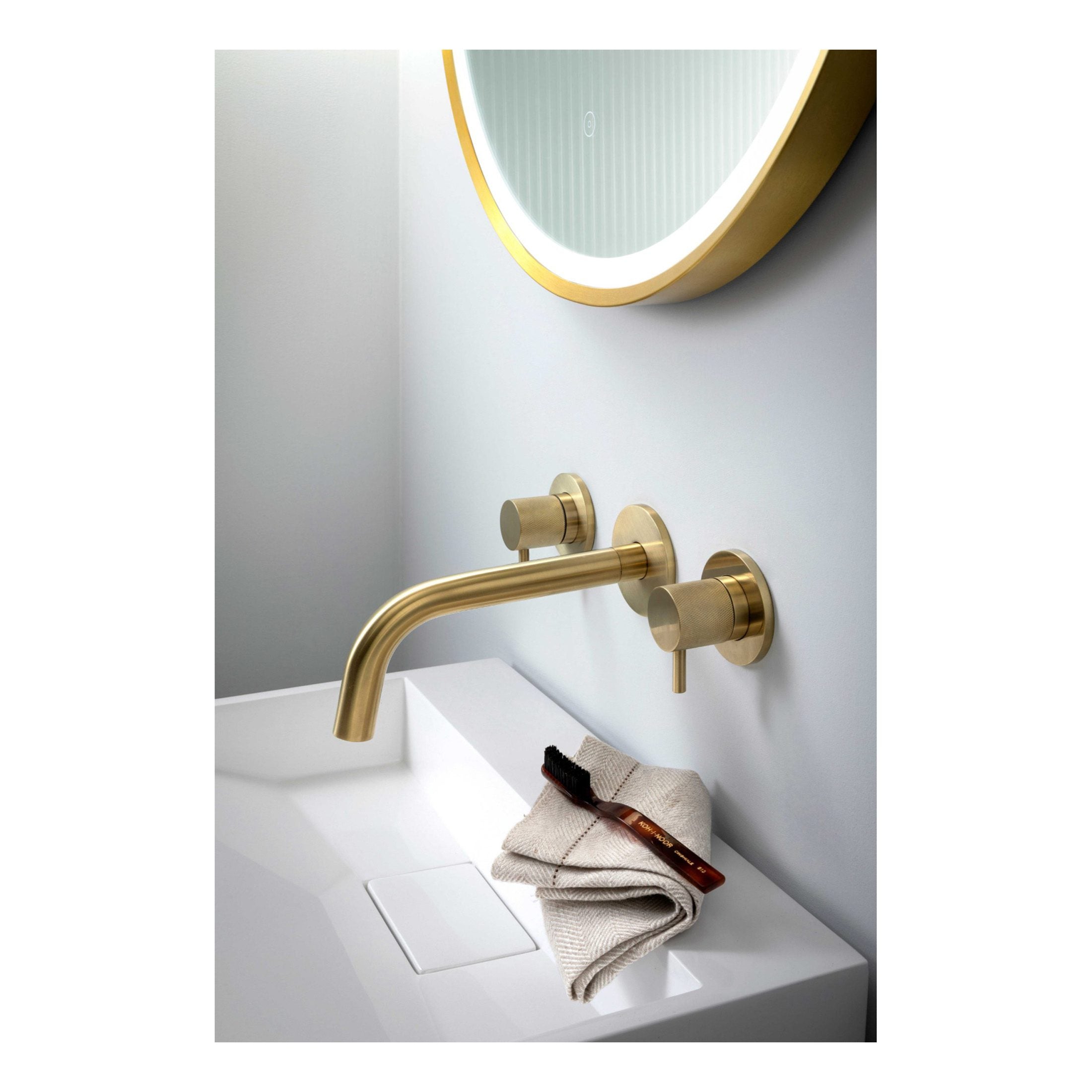 Sleek gold bathroom faucet with wall-mounted controls above a modern white sink, complemented by round illuminated mirror, showcasing elegant bathroom fixtures for luxurious Bubly Bathrooms.