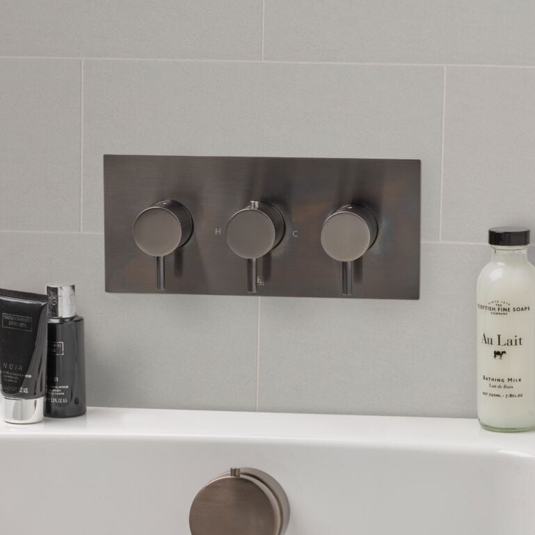 Sleek modern bathroom thermostatic shower control with three stainless steel dials, set against light grey tiled wall. Includes bath products on the white ledge beneath.