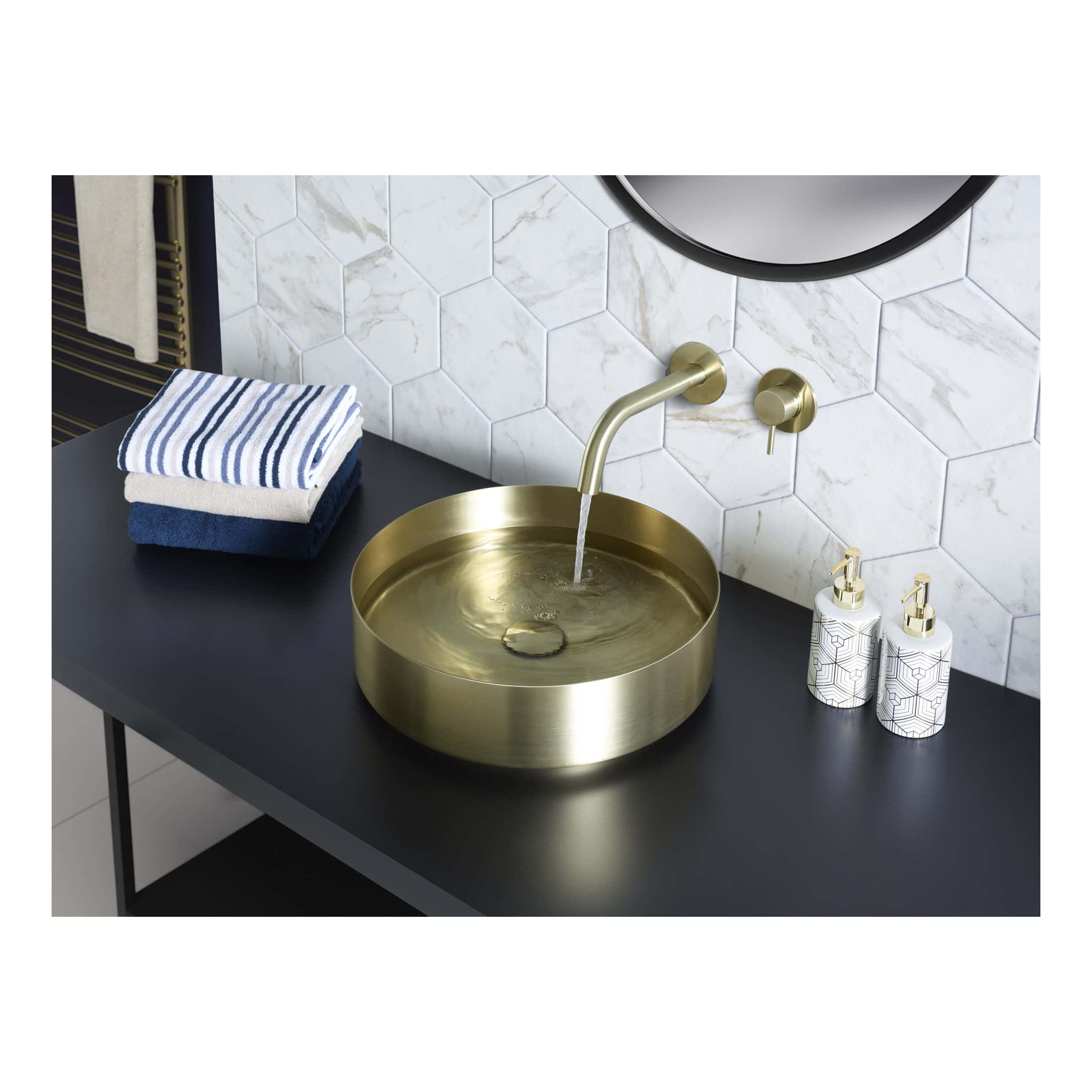 Luxurious round brass bathroom sink with modern wall-mounted tap, set against a white marble hexagon tile wall, complemented by neatly folded towels and stylish soap dispensers, perfect for a contemporary bathroom design.