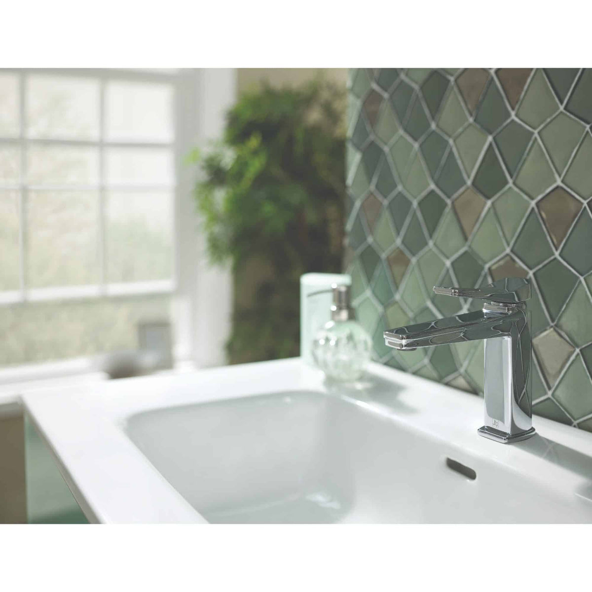Shiny chrome bathroom faucet on a white sink with decorative green tile backsplash, modern bathroom design, natural light from large window, stylish and functional bathroom fixtures.