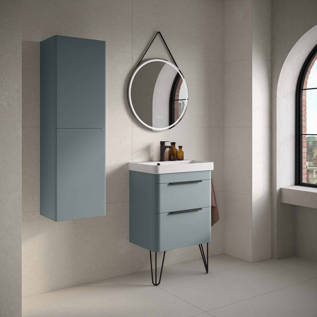 Sleek blue bathroom vanity unit with modern black handles and tripod style legs, accompanied by a matching vertical storage cabinet, under a round illuminated mirror in a stylish, contemporary bathroom setting.