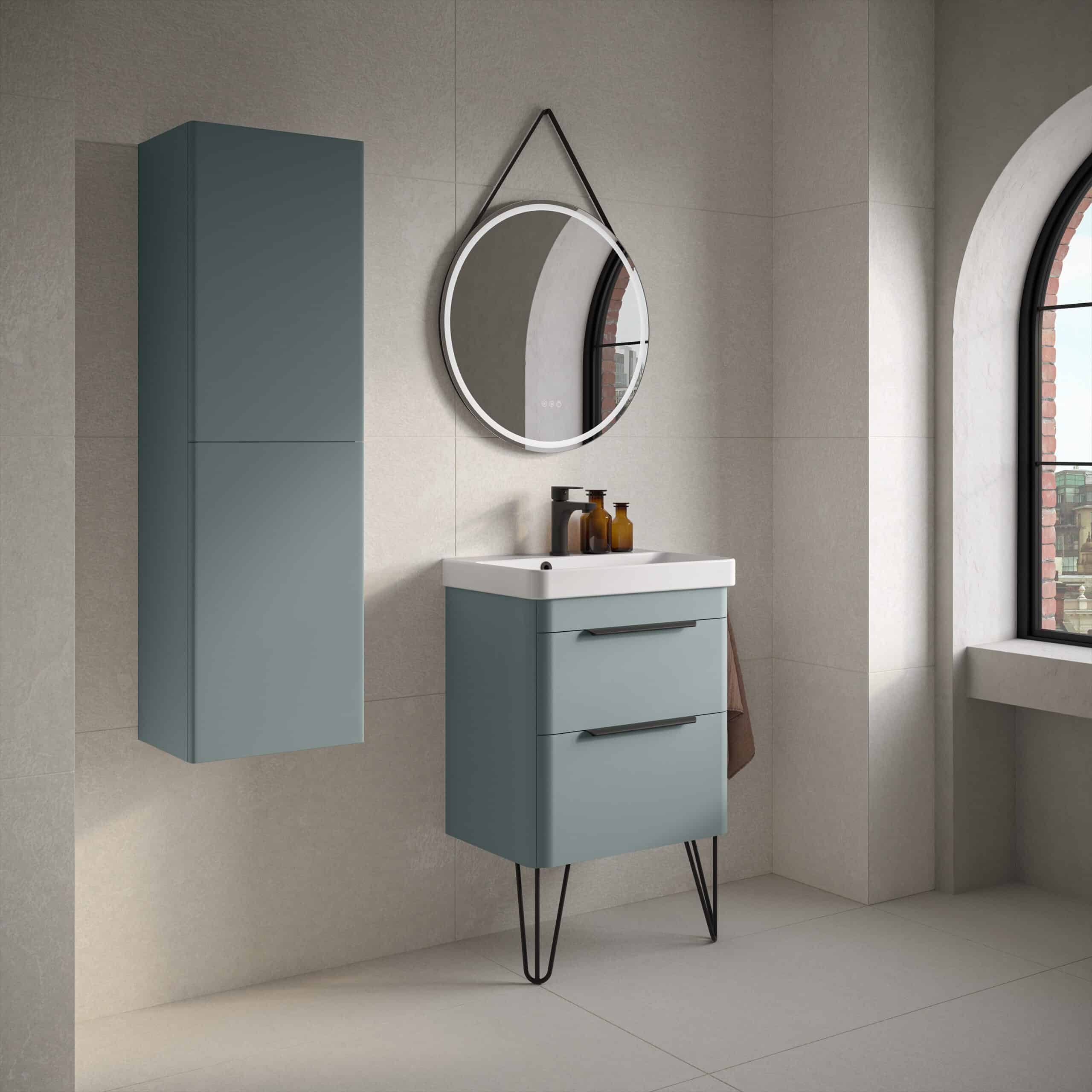 Sleek blue bathroom vanity unit with modern black handles and tripod style legs, accompanied by a matching vertical storage cabinet, under a round illuminated mirror in a stylish, contemporary bathroom setting.