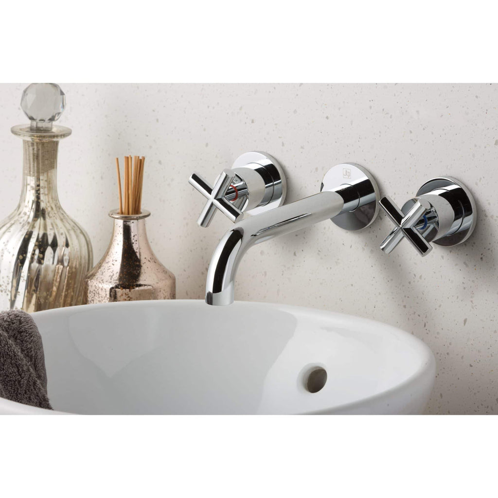 Polished chrome wall-mounted bathroom tap with twin cross handles for hot and cold water, installed above a white ceramic sink with decorative bottles and diffusers in a modern bathroom setting.