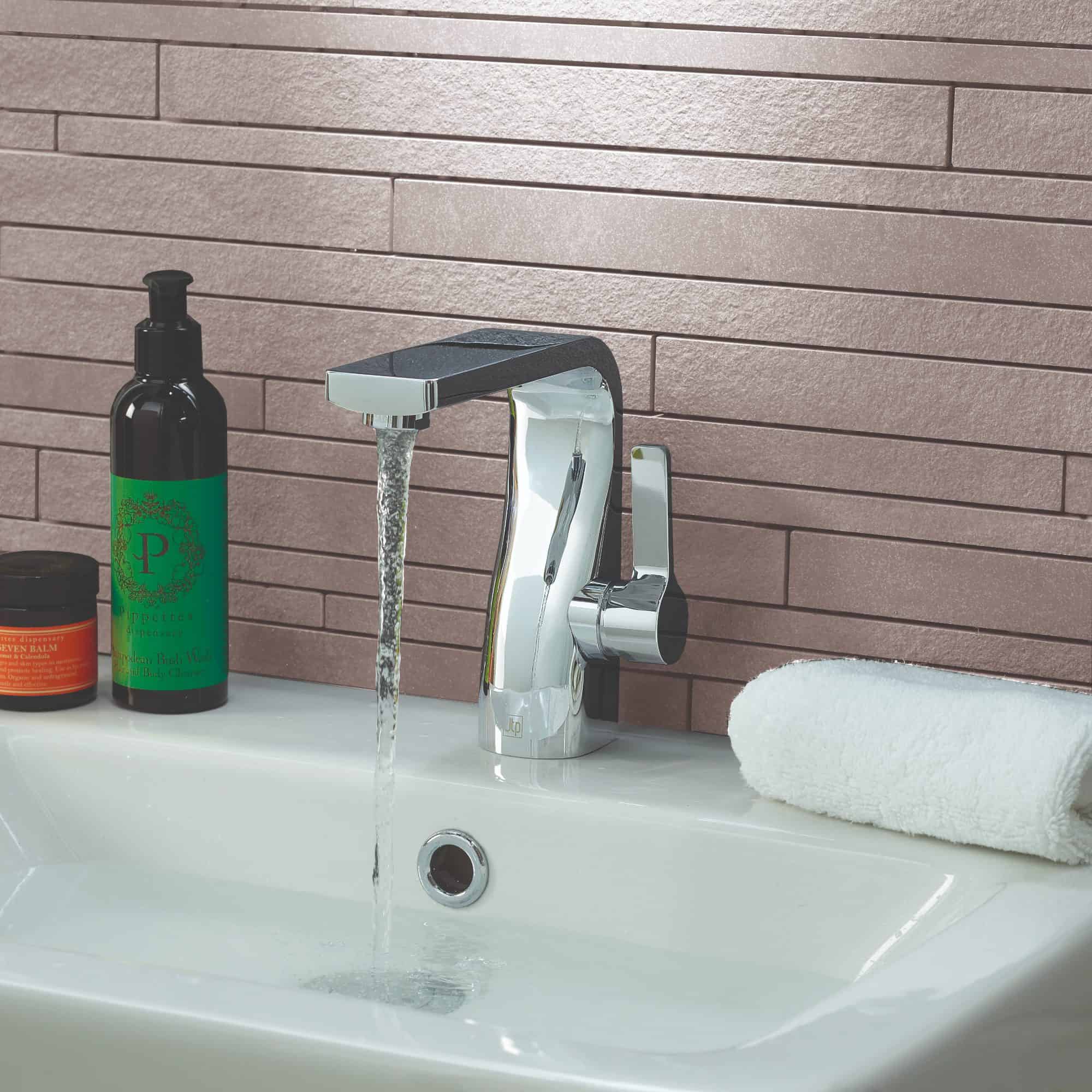 Sleek modern chrome bathroom tap with running water, white sink, rolled towel, and bathroom essentials in the background.