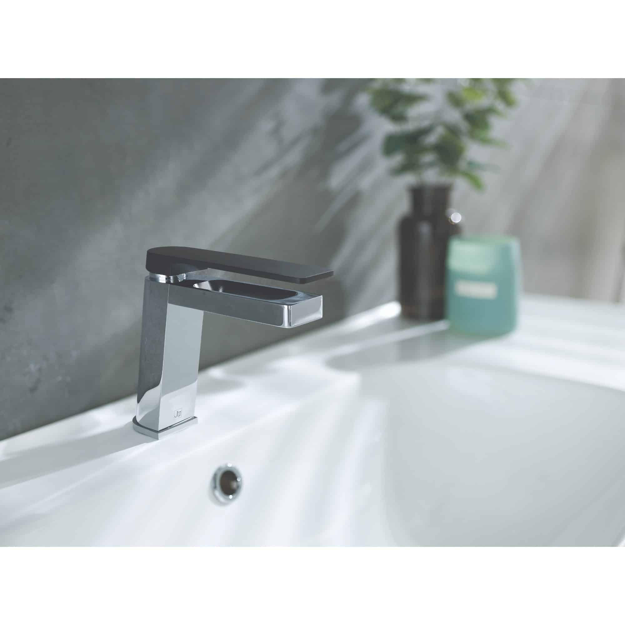 Sleek chrome bathroom sink tap with modern design, installed on a white ceramic basin, with decorative bottles and a plant in the background, ideal for contemporary bathroom decor.