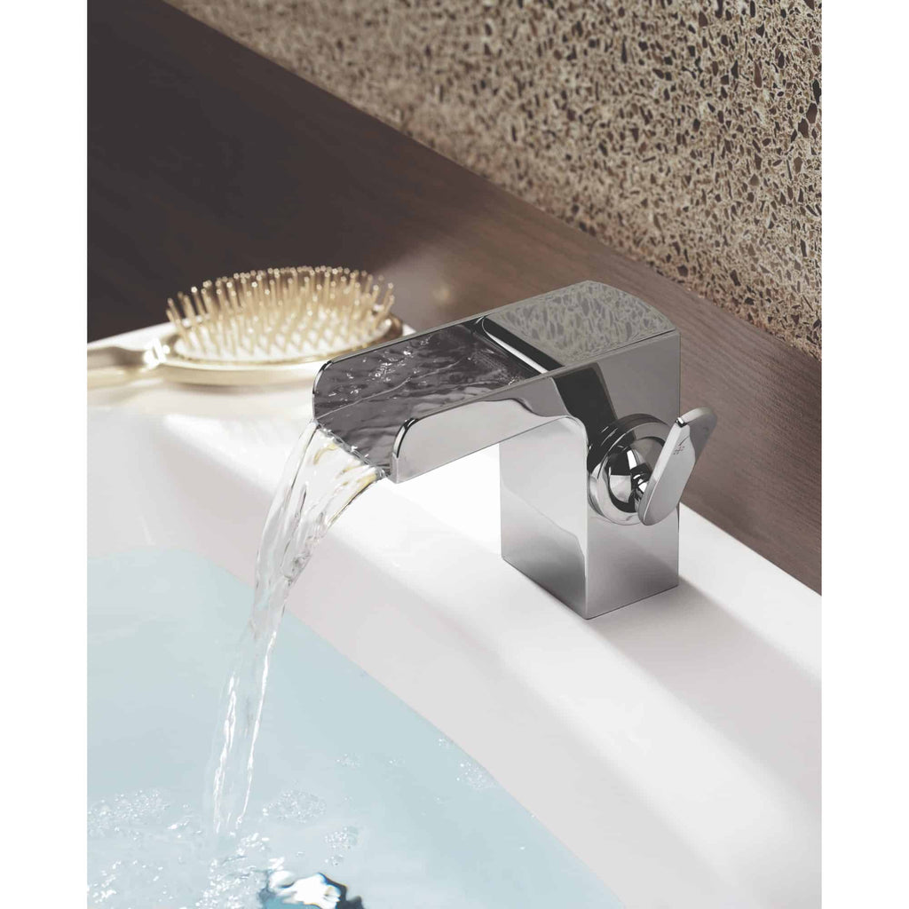 Sleek modern chrome bathroom tap with water flowing into a white bathtub, set against textured brown wall and wooden shelf, exemplifying contemporary bathroom design and fixtures.