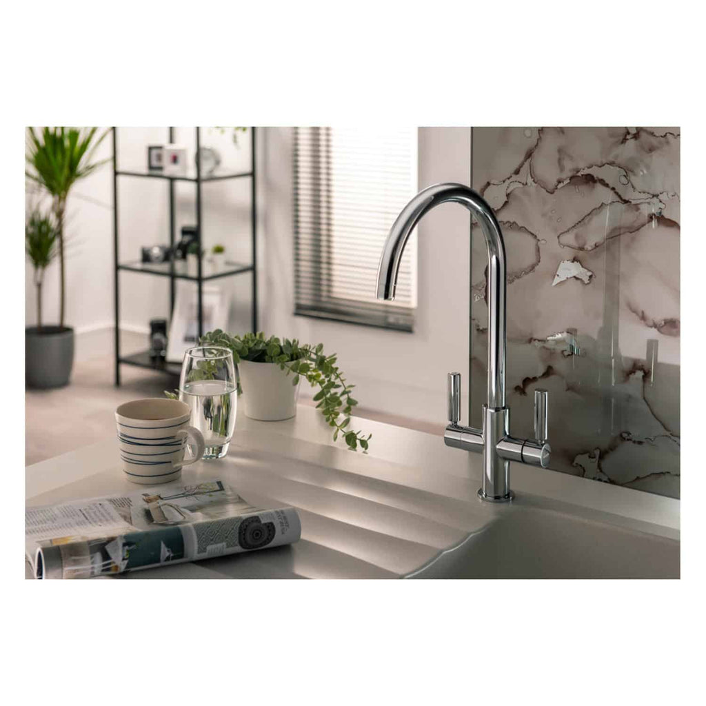 Sleek chrome kitchen sink tap with a modern design, installed on a white countertop with a marble-style backsplash, surrounded by a potted plant, cup, glass of water, and a magazine.