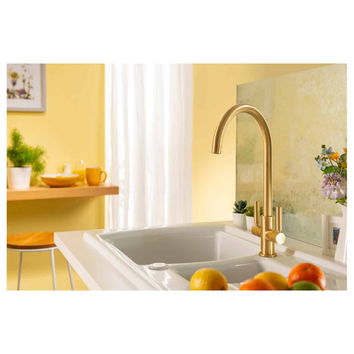 Elegant kitchen sink with a modern gold faucet, surrounded by fresh fruits and decorative plants in a bright, contemporary kitchen with yellow walls and white curtains.