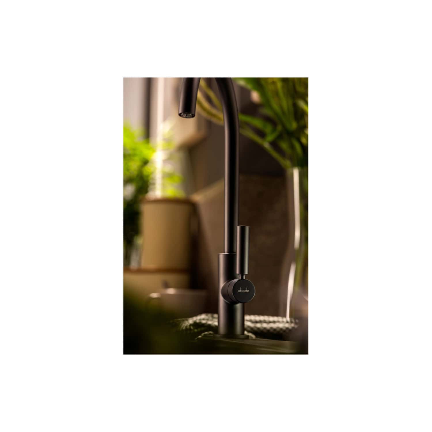 Sleek black modern kitchen tap with a pull-out spray feature, branded "abode", installed in a contemporary kitchen setting with natural light and lush green plants in the background.