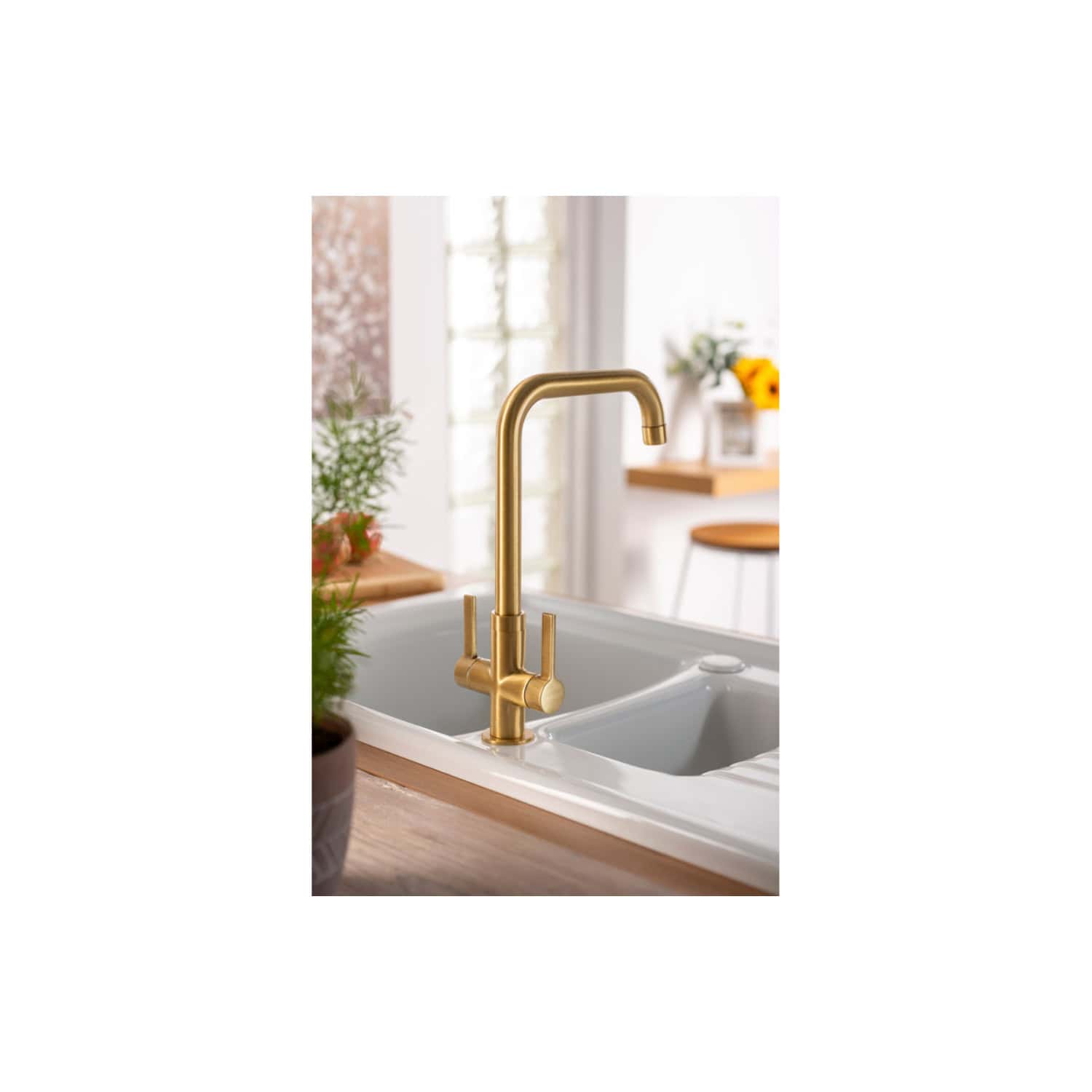 Brass kitchen sink tap with modern design, installed in a white ceramic sink in a bright kitchen with natural light, wood accents, and decorative plants for a stylish, functional space.