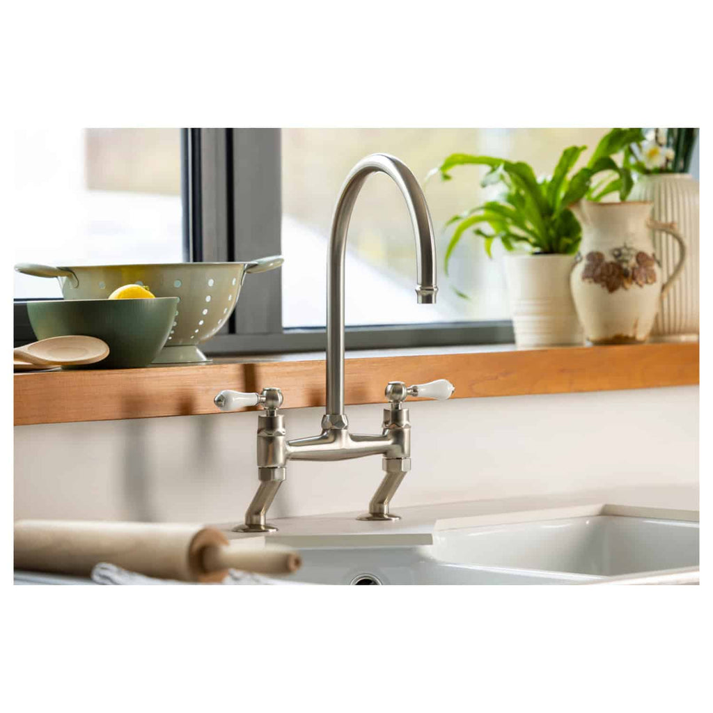Polished stainless steel kitchen sink mixer tap with dual handles, modern design, installed in a contemporary kitchen with wooden countertop and decorative plants.