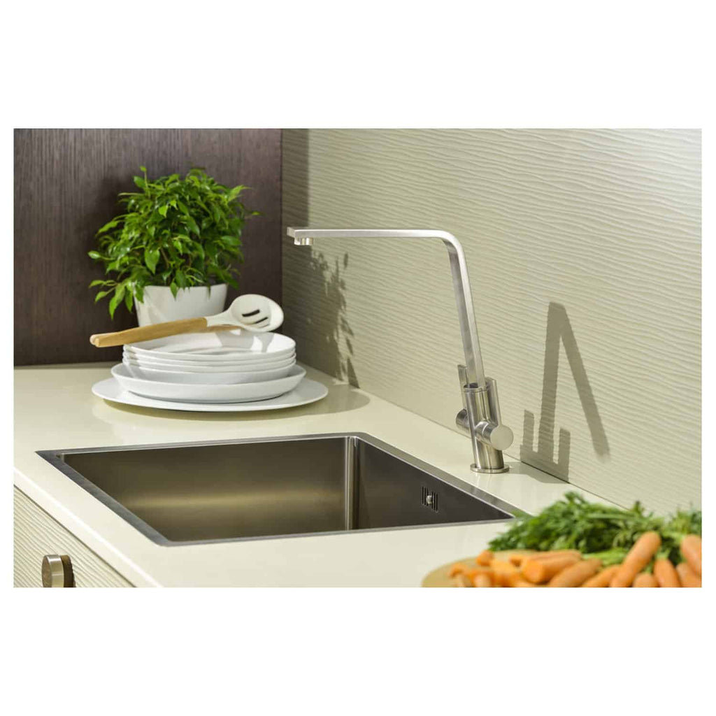 Modern stainless steel kitchen sink with sleek tap, surrounded by white plates, green plants, and fresh carrots, showcasing contemporary design and practical functionality.