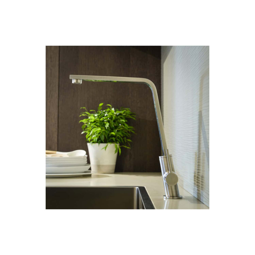 Brushed stainless steel kitchen tap with a modern curved design, installed on a beige countertop near a window, with a white ceramic potted plant and stacked white plates in the background, showcasing contemporary kitchen fixtures and decor.