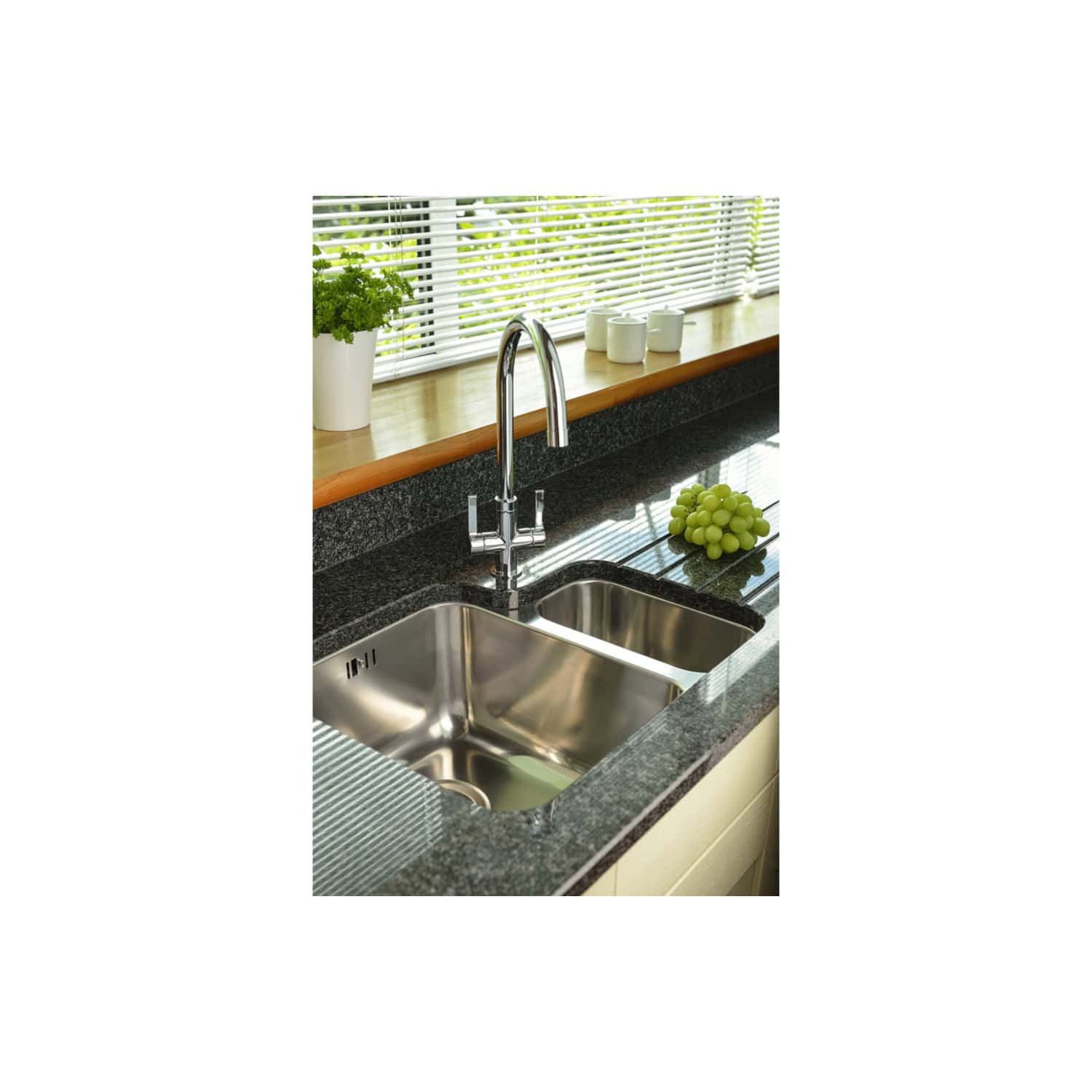 Stainless steel kitchen sink with chrome faucet, black granite countertop, window with blinds, green grapes, and white mugs on wooden windowsill in modern bathroom or kitchen setting.