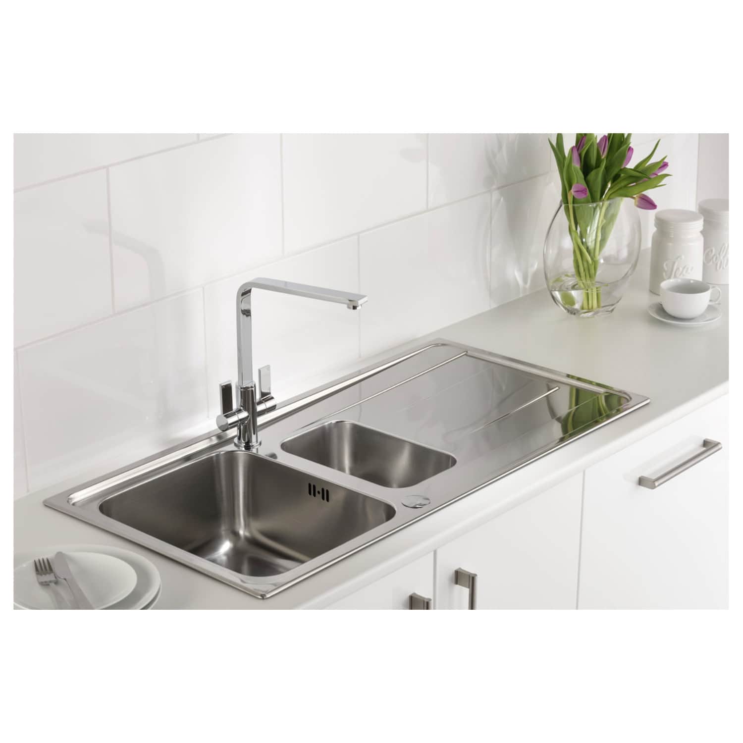 Stainless steel double kitchen sink with modern tap, set in a white kitchen with tiled wall, alongside a vase of purple tulips and kitchen canisters, offering stylish and functional kitchen design.