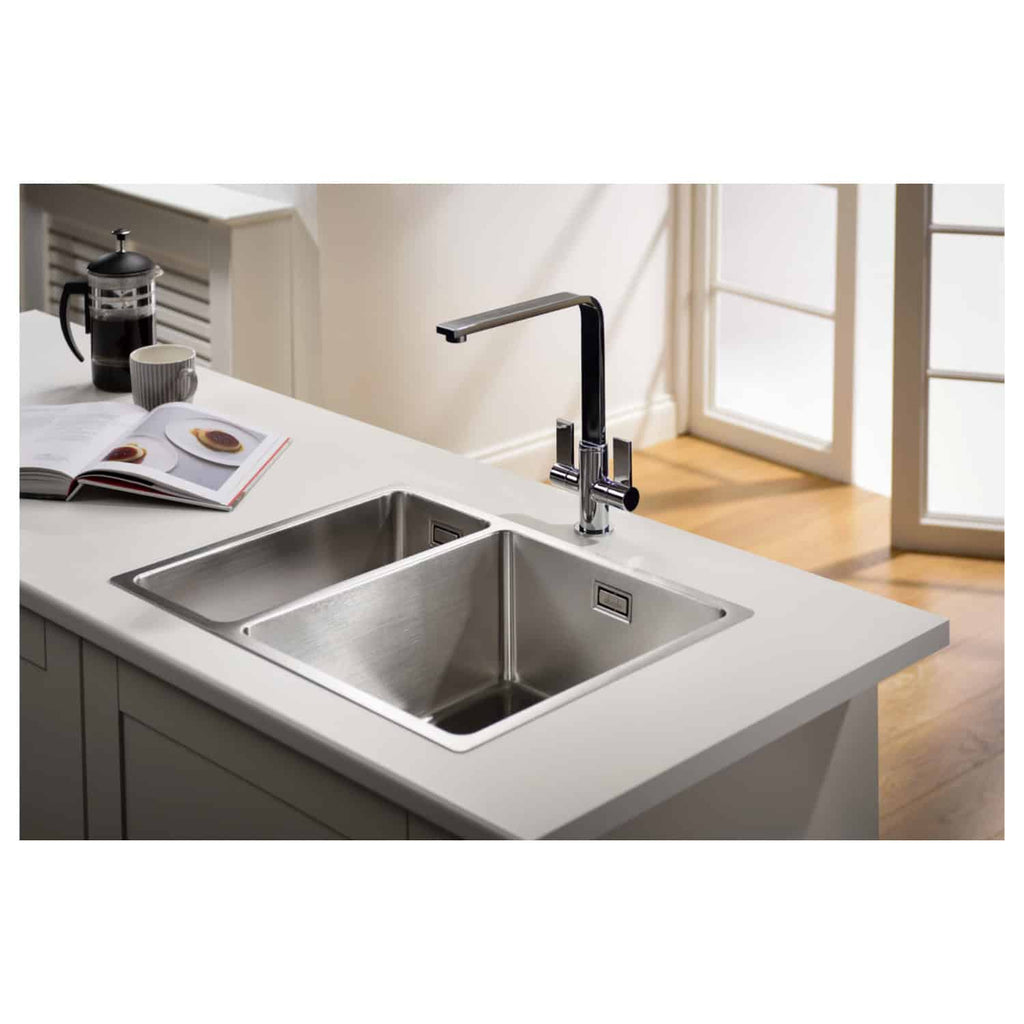Sleek stainless steel kitchen sink with modern chrome faucet, integrated into a white countertop, in a bright contemporary kitchen with wooden flooring and natural light from glass doors.