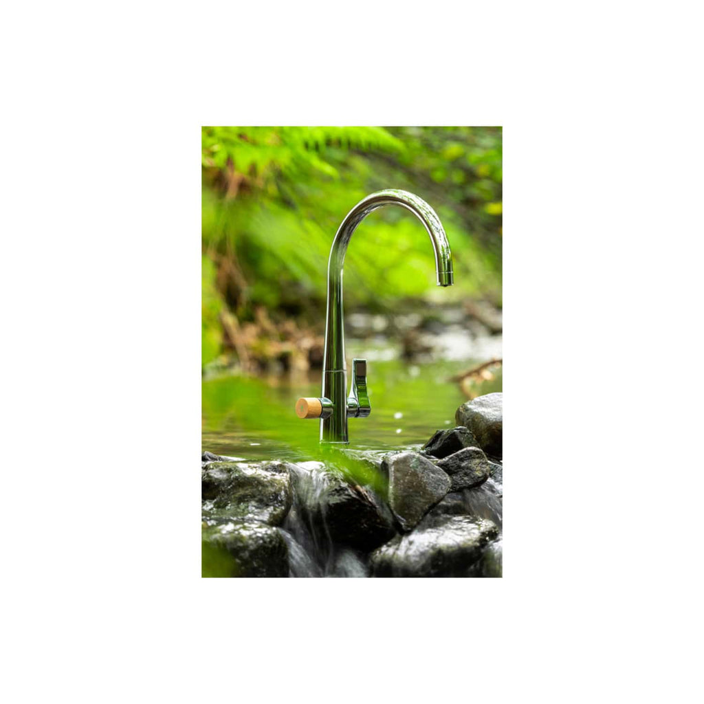 Futuristic outdoor brass bathroom faucet mounted on rocks near a flowing stream with lush green foliage in the background, showcasing eco-friendly outdoor plumbing solutions.