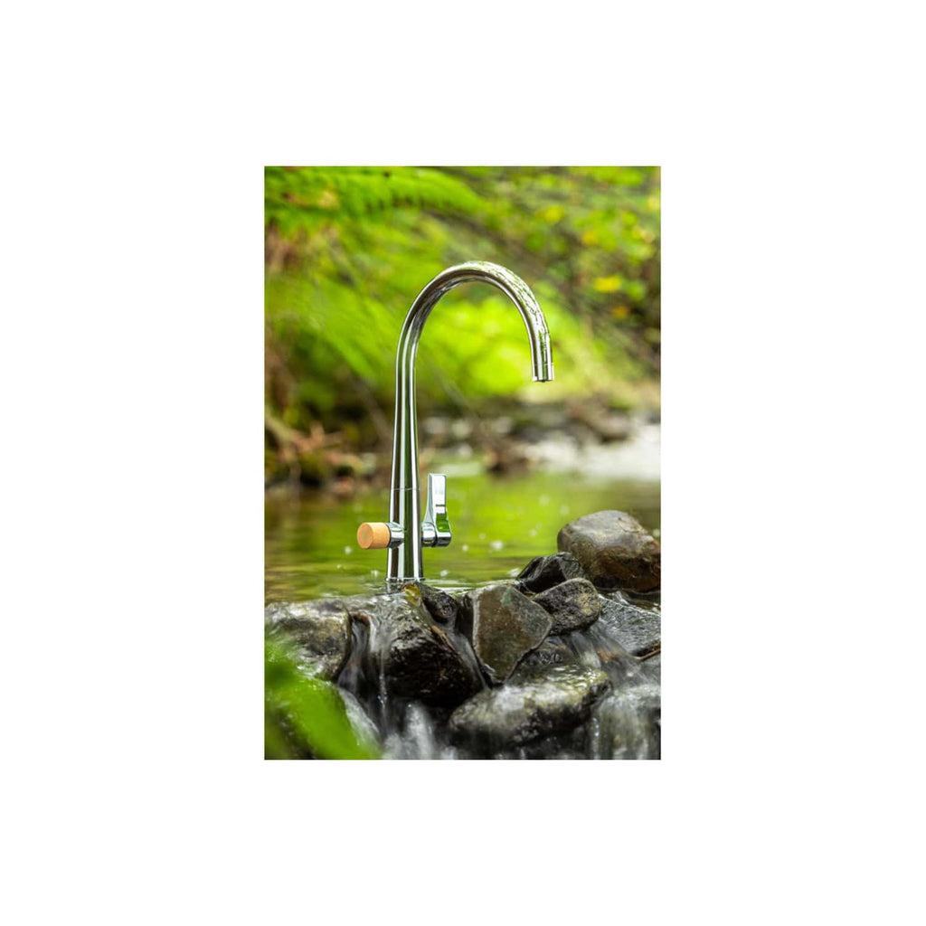 Shiny outdoor kitchen tap with wooden handle installed above rocks in a natural stream, surrounded by lush green foliage. Perfect for outdoor bathroom or garden water feature.