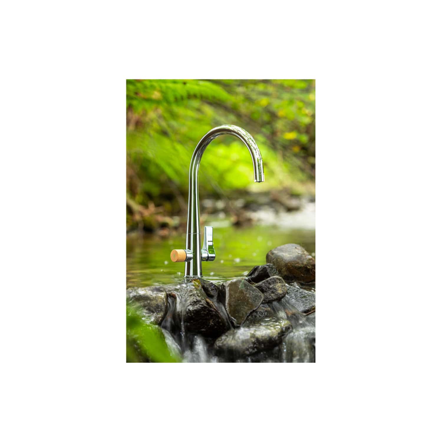 Shiny outdoor kitchen tap with wooden handle installed above rocks in a natural stream, surrounded by lush green foliage. Perfect for outdoor bathroom or garden water feature.