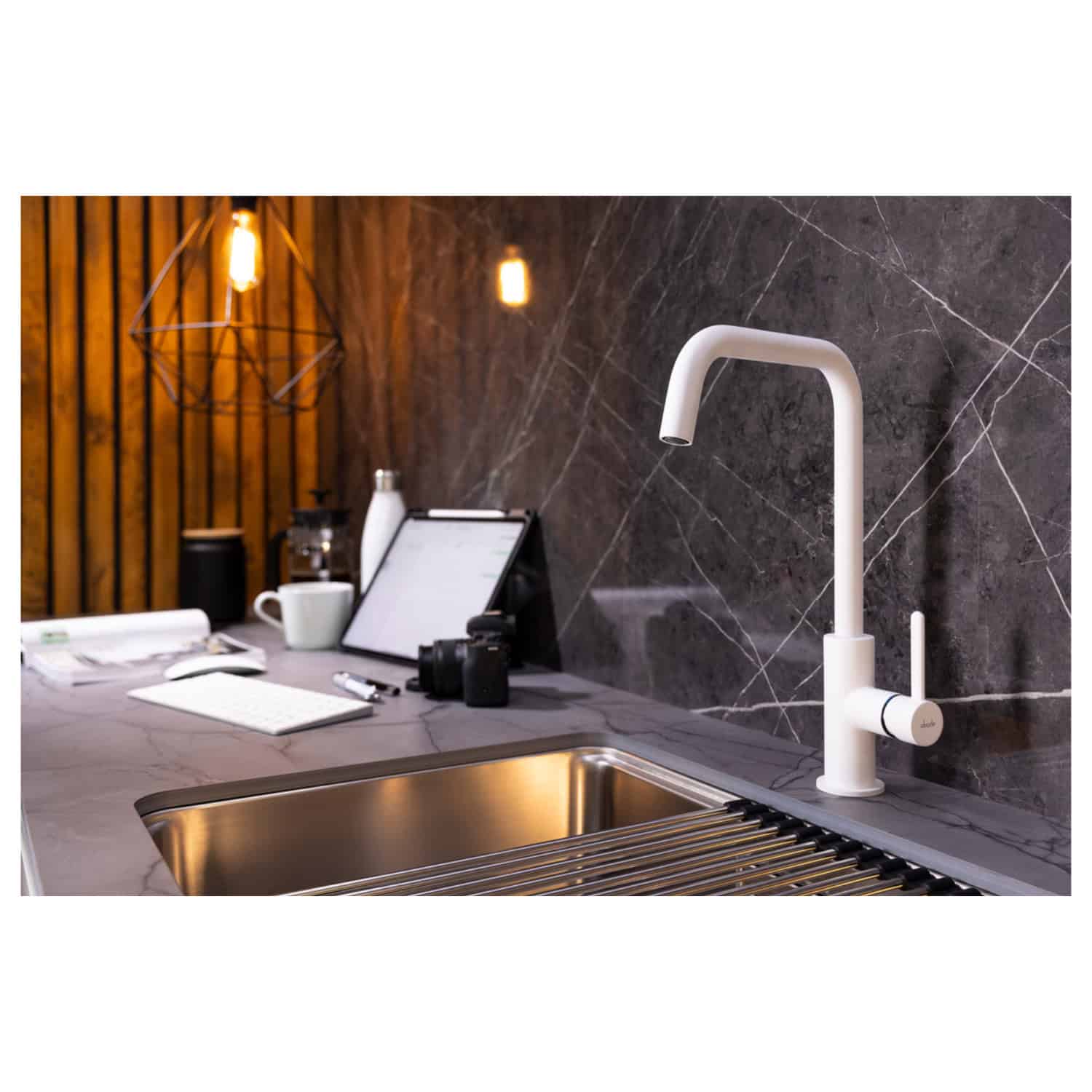 Sleek white kitchen tap installed above a stainless steel sink, with modern decor and wooden wall accents in the background. The setup includes a tablet, camera, and other kitchen essentials, highlighting contemporary design.