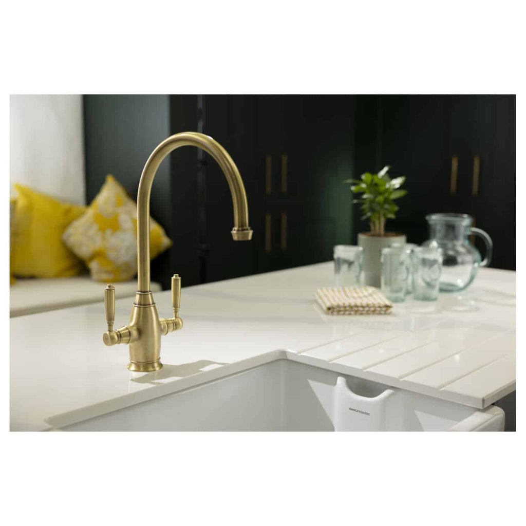 Sleek brass kitchen tap over a modern white sink with a stylish kitchen countertop, complemented by minimal decor including glassware, a small plant, and a black cabinet in the background.