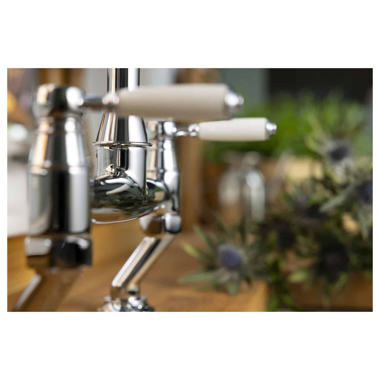 Shiny chrome bathroom tap with white handles on wooden countertop, with blurred floral decor in the background, showcasing modern bathroom fixtures.