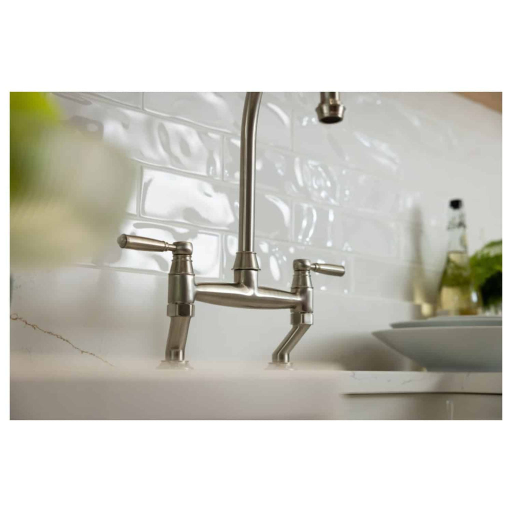Sleek stainless steel kitchen tap with dual handles, mounted on a white marble countertop, set against a glossy white tile backsplash in a modern bathroom or kitchen setting.