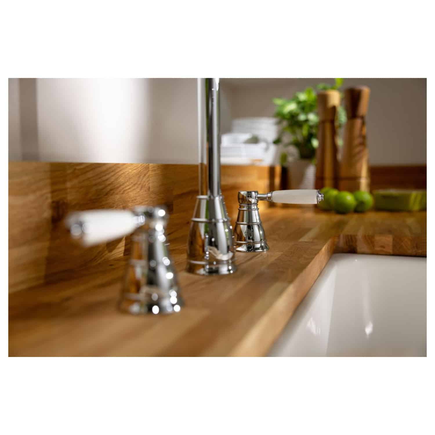 Polished chrome bathroom tapware with white ceramic handles on a wooden vanity, featuring a large mirror, green limes, and kitchen accessories in the background, showcasing elegant bathroom design.