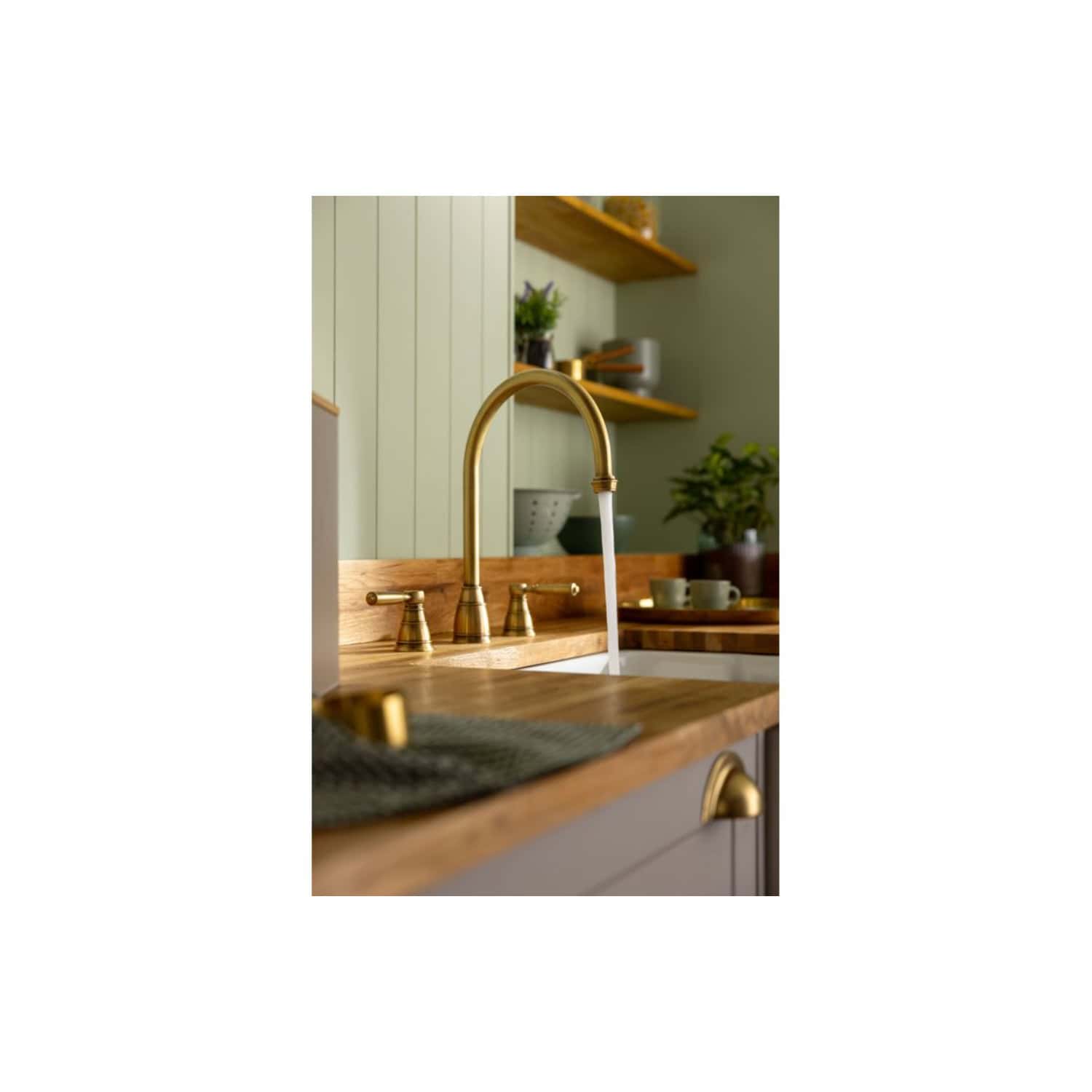 Sleek brass kitchen tap with flowing water set against a contemporary farmhouse kitchen design, featuring wooden countertops, green cabinets, and decorative shelves with plants and ceramics.
