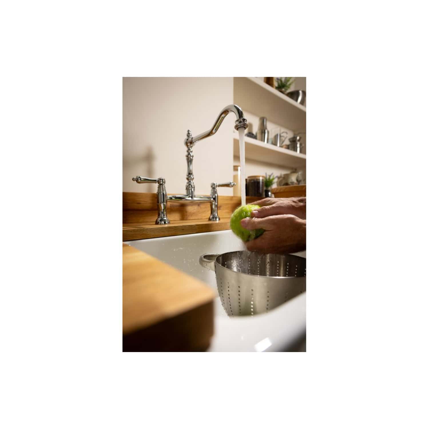 Clean kitchen sink with chrome tap, hand washing a green apple, stainless steel colander, and wooden countertop, showcasing contemporary bathroom fixtures and accessories.