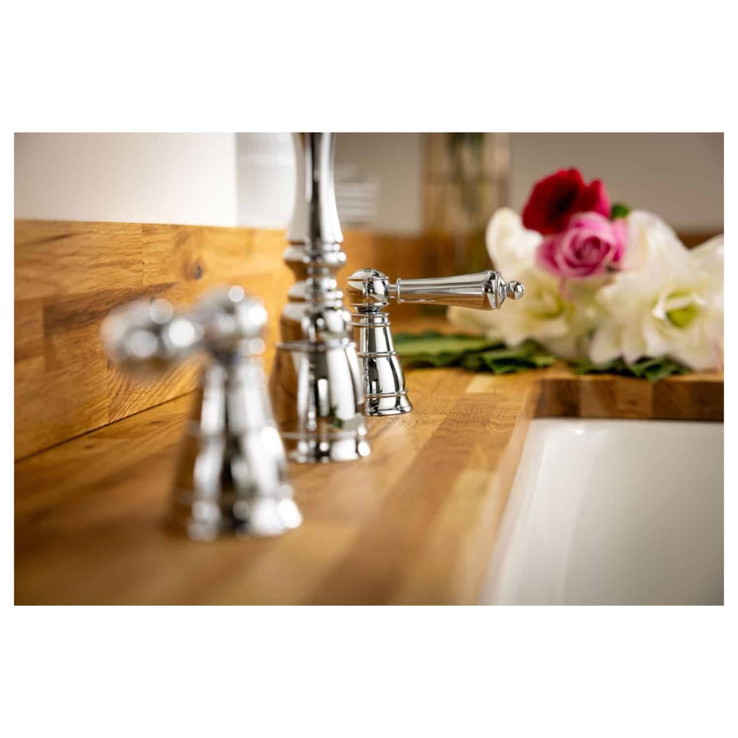 Shiny chrome bathroom faucet set with a wooden countertop and elegant floral arrangement in the background, showcasing luxury bathroom fixtures and decor.