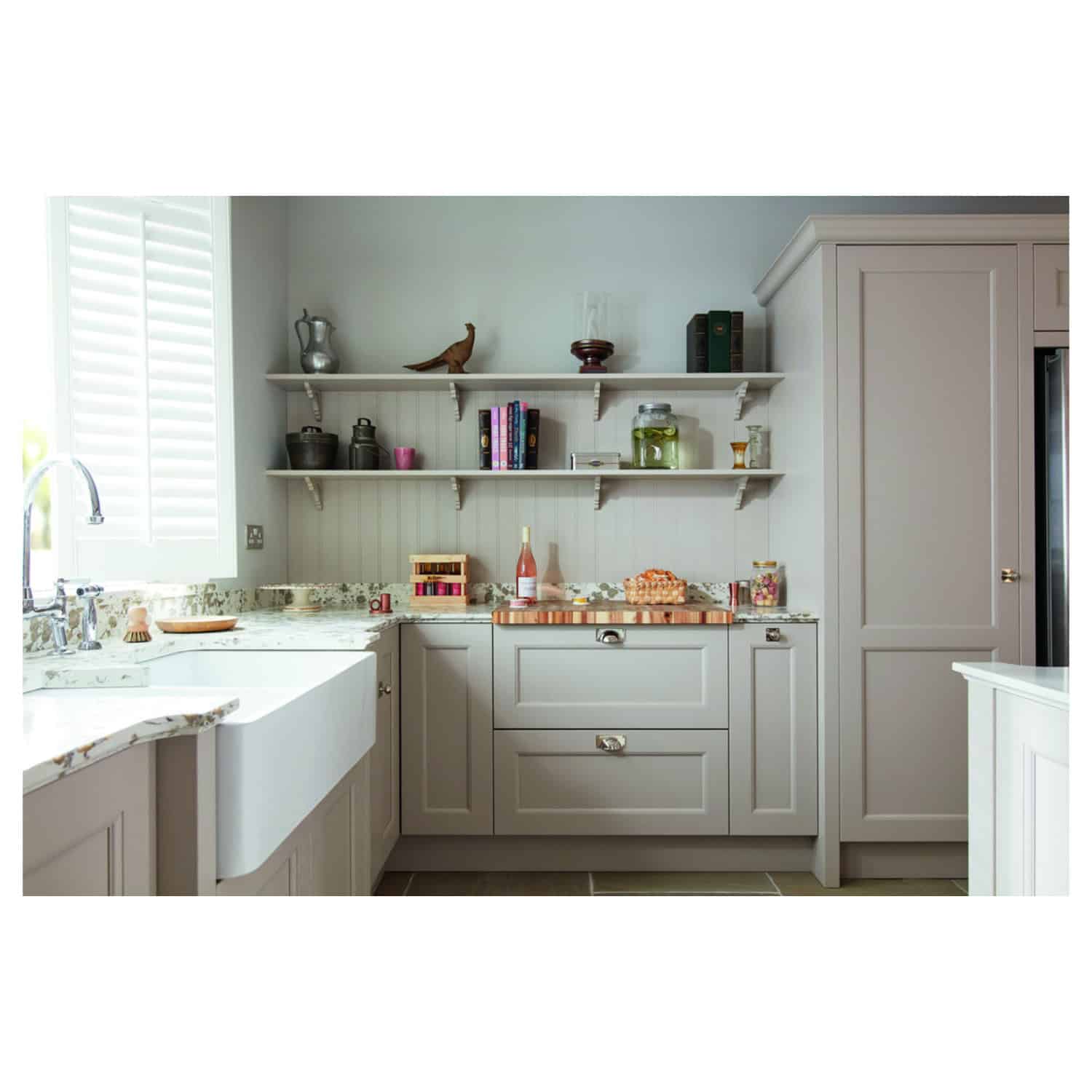 Streamlined grey kitchen cabinets with marble countertops, open shelving displaying cookbooks and decorative items, bright natural light from window with white shutters, modern and functional kitchen design.