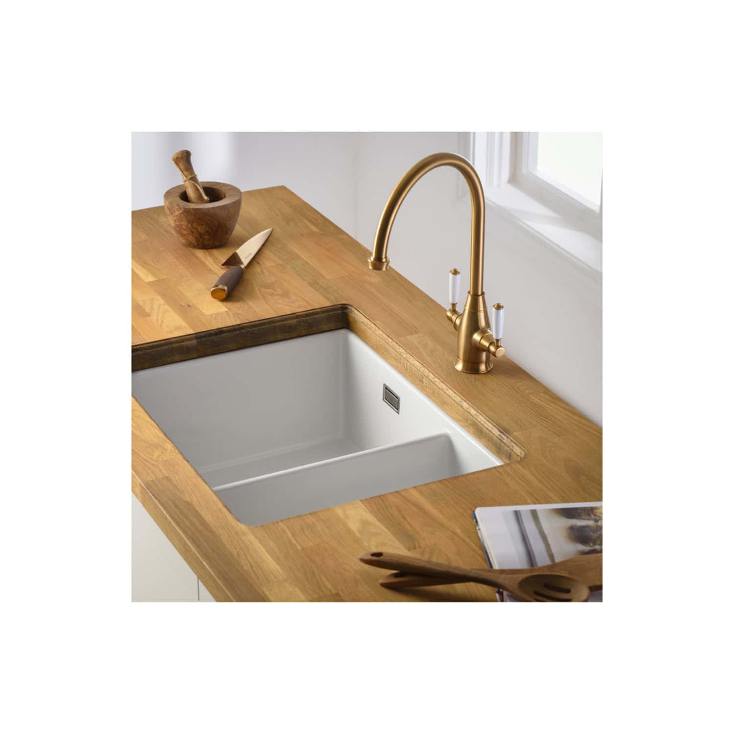 Polished brass kitchen tap with a deep white farmhouse sink on a wooden countertop, modern kitchen design with natural light.