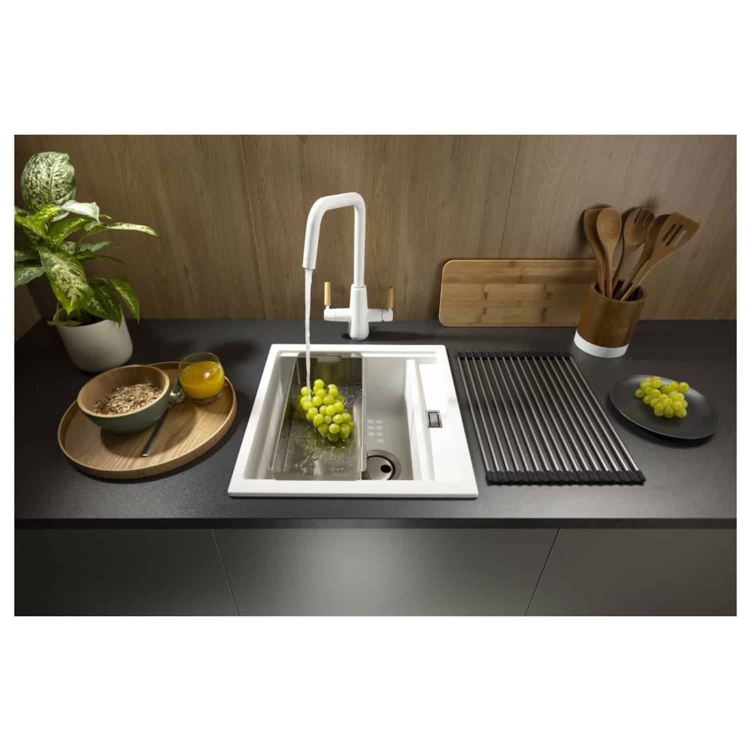 Modern kitchen sink with white matte finish and pull-out spray faucet set against a wooden backsplash, surrounded by fresh grapes, wooden utensils, cereal, and juice, highlighting stylish and functional kitchen design.