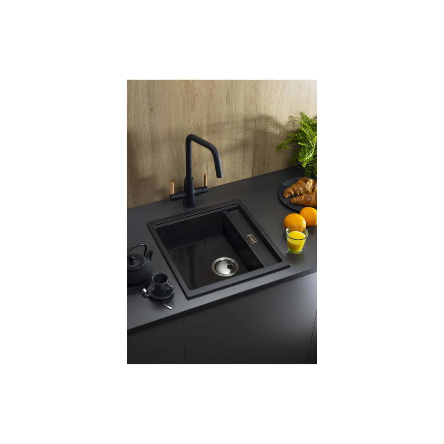 Sleek black kitchen sink with modern black faucet, surrounded by fresh oranges, ginger, a glass of orange juice, and a green plant on a dark countertop with a wood backdrop, showcasing stylish kitchen design.