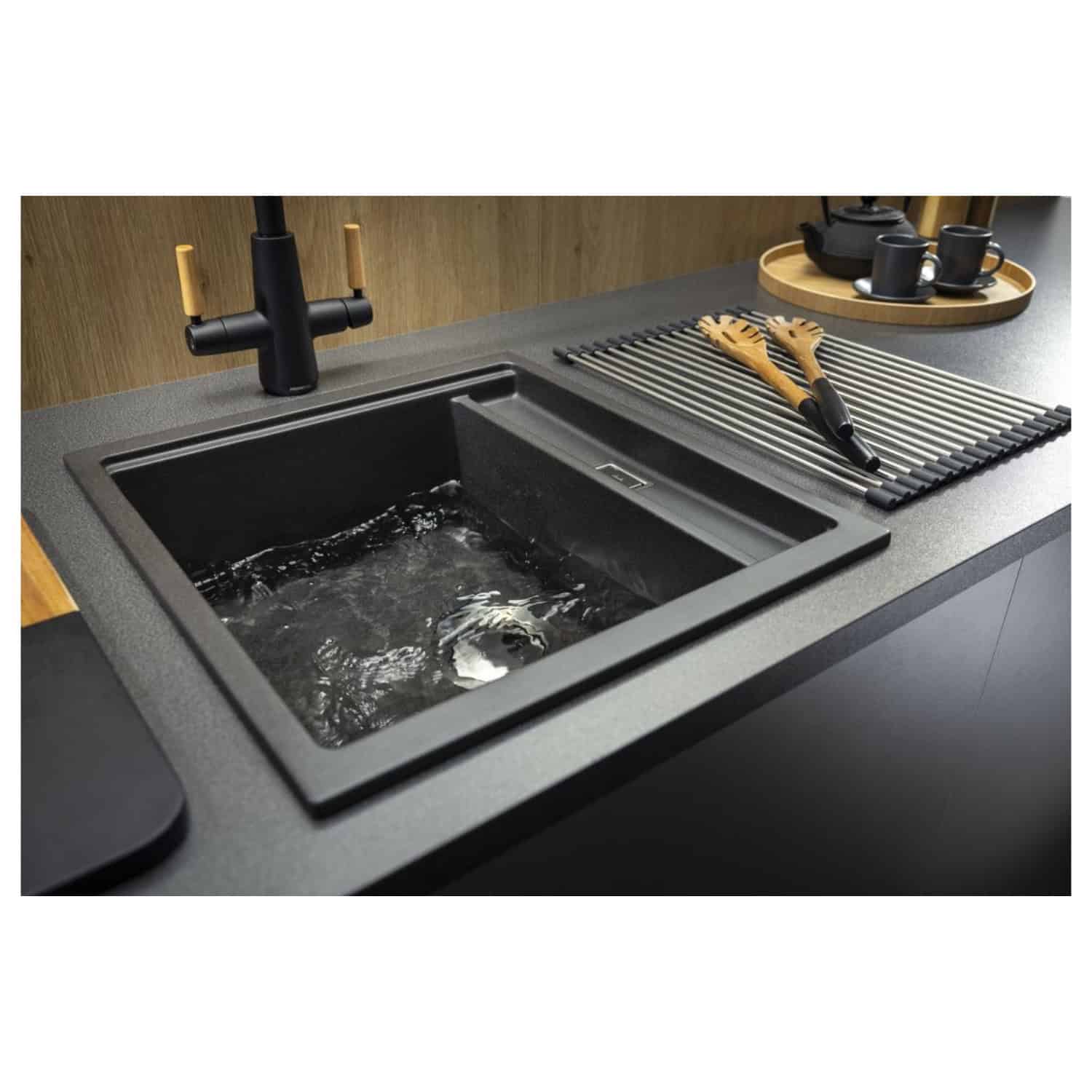 Black kitchen sink with water running, modern matte black faucet, wooden utensils, drying rack, and black tea set on countertop - Bubly Bathrooms.