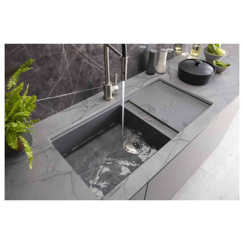 Shallow stainless steel kitchen sink with integrated draining area and modern chrome faucet, set in a grey marble countertop, with green plants and black cookware for a sleek bathroom or kitchen design.