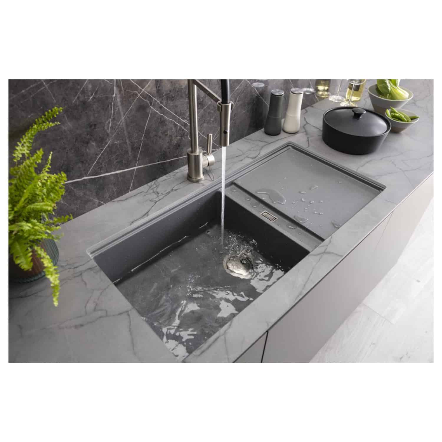 Shallow stainless steel kitchen sink with integrated draining area and modern chrome faucet, set in a grey marble countertop, with green plants and black cookware for a sleek bathroom or kitchen design.