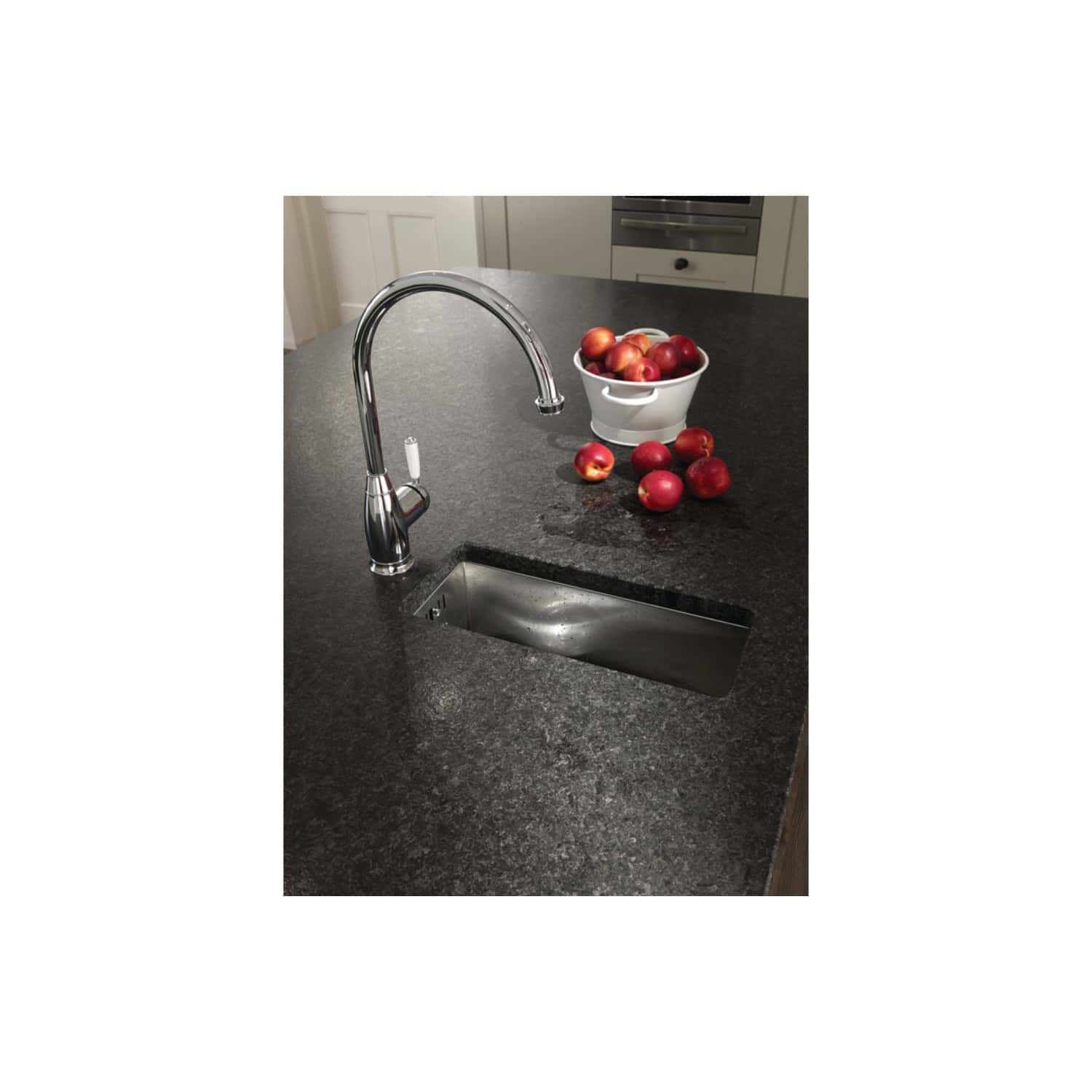 Shiny chrome kitchen sink faucet with a modern black granite countertop, next to a white bowl filled with fresh red and yellow apples in a contemporary kitchen setting.