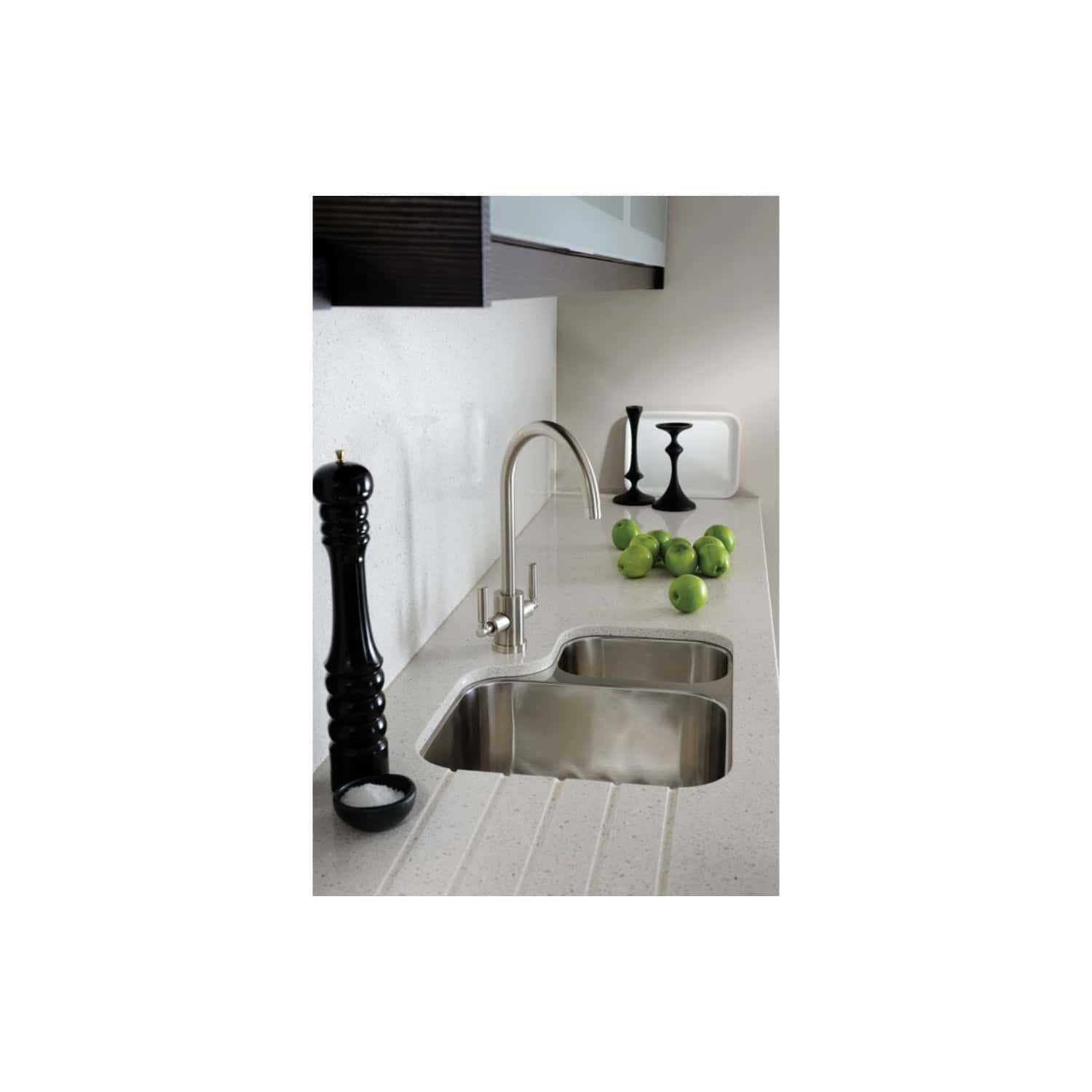 Shiny stainless steel kitchen sink with modern fixtures, black pepper grinder, green apples, and sleek black candlesticks on a light-colored countertop in a contemporary kitchen design.
