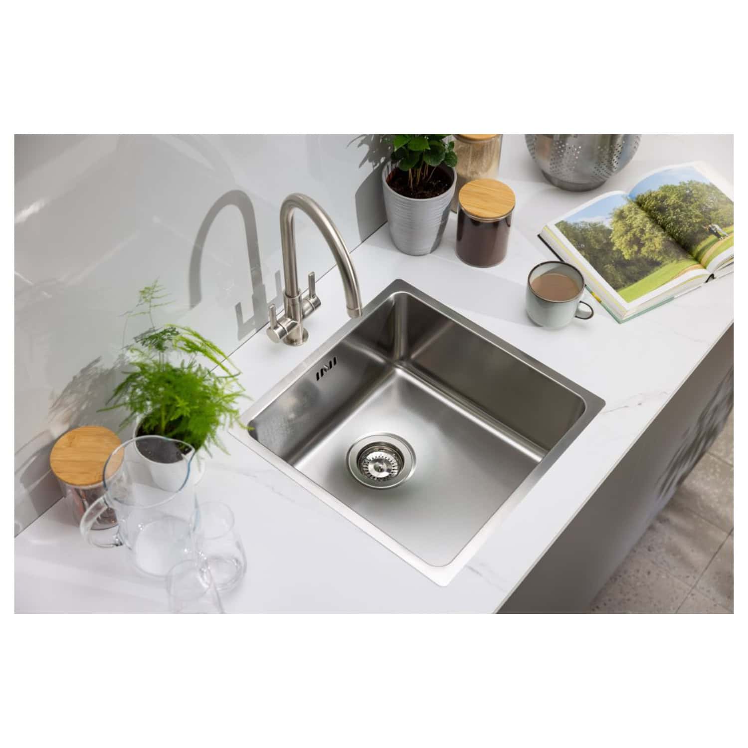 Shiny stainless steel kitchen sink with modern chrome tap, surrounded by decorative plants, glassware, a cup of tea, an open magazine, and stylish storage jars on a white marble countertop in a contemporary bathroom setting.