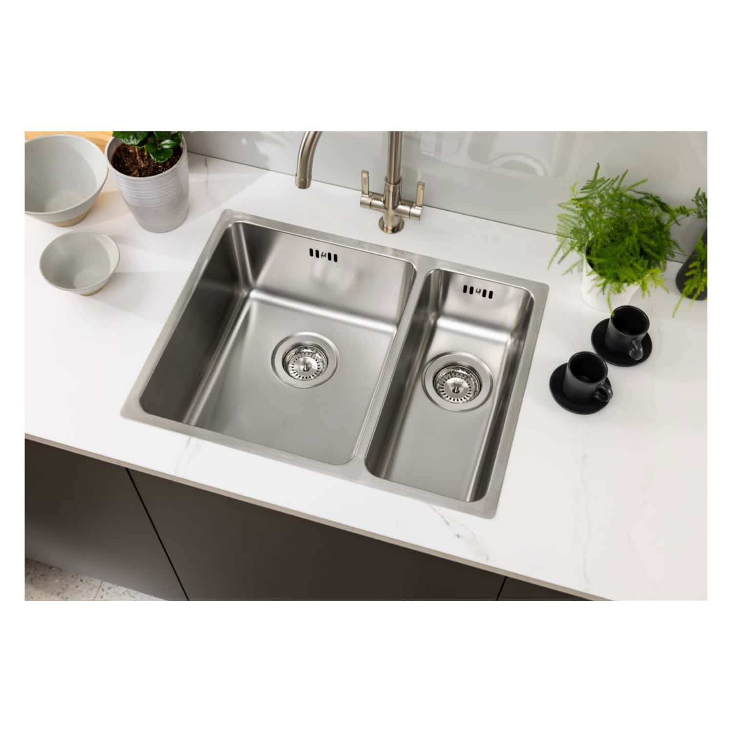 Stainless steel kitchen sink with dual bowls, modern design, fitted in a sleek white countertop with decorative black cups, green plants, and minimalistic kitchen accessories.